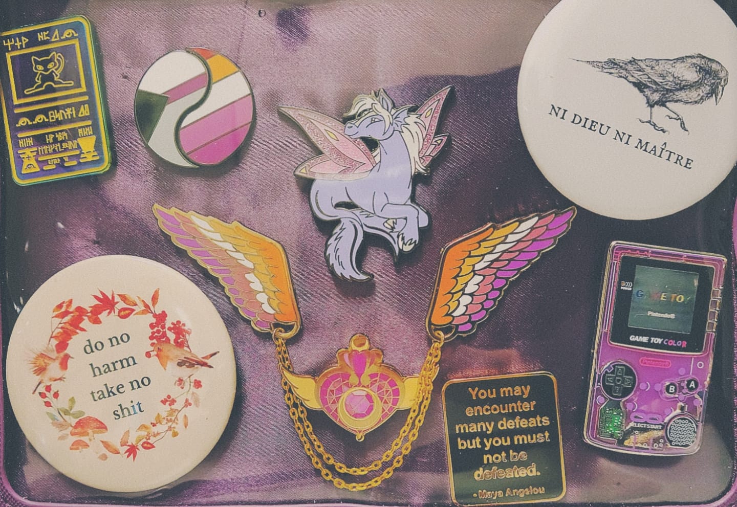 Kyr's ita bag. It is purple and the pin pad has a satin exterior.  Pins include a chrome Mew pin, demisexual and lesbian pride flag yin yang pins, an unconverted Faerie Peopin, a crow captioned 'Ni Dieu Ni Maître', the quote 'do no harm take no shit' inside an autumn colored wreath with two small robin birds and mushrooms, lesbian pride themed angel wings, a Crisis Moon Compact, the quote 'You may encounter many defeats but you must not be defeated' by Maya Angelou, and a translucent purple Game Boy Color.